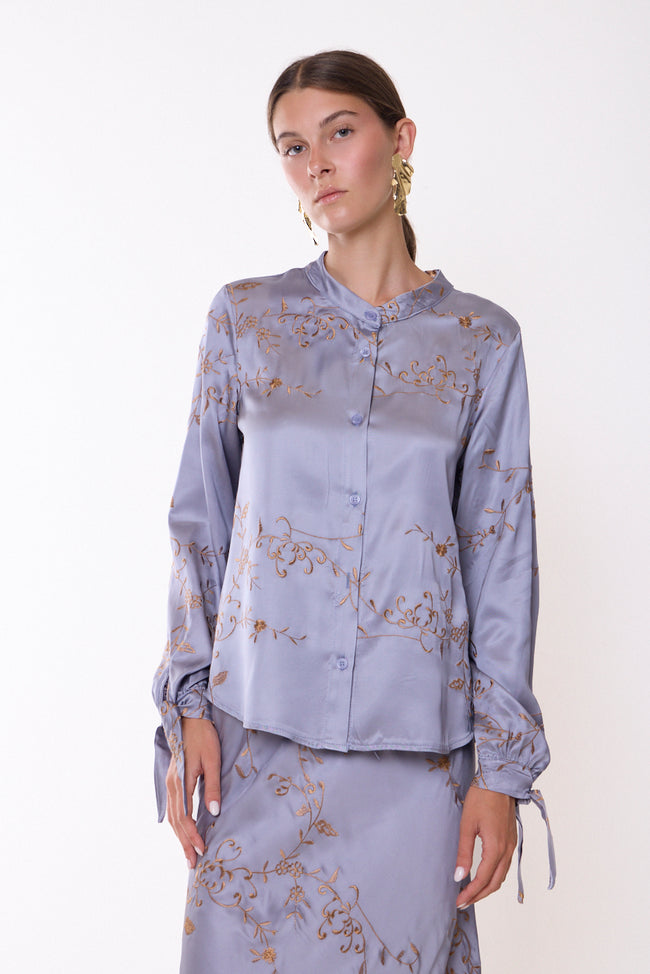 Grey lilac-toned band-collar button-front blouse with floral vine and scrolling embroidery, long sleeves and cuff ties; coordinating skirt and shoes styled separately