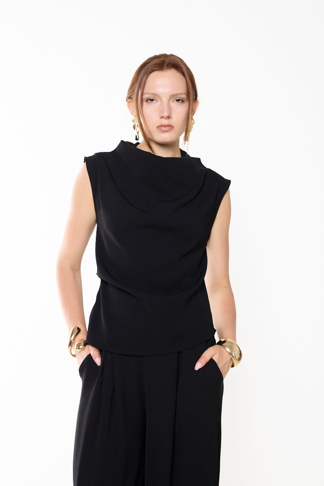 Black draped high-neck sleeveless top with asymmetric front overlay and shaped silhouette; wide-leg trousers and gold-tone accessories styled separately
