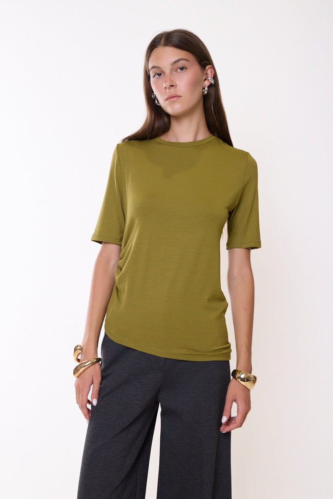 Olive green round-neck short-sleeve T-shirt with side gathers and asymmetric hem; wide-leg trousers and jewellery styled separately