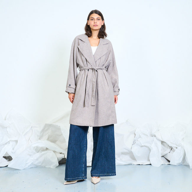 Model wearing a light neutral belted coat with notched lapels and relaxed sleeves.