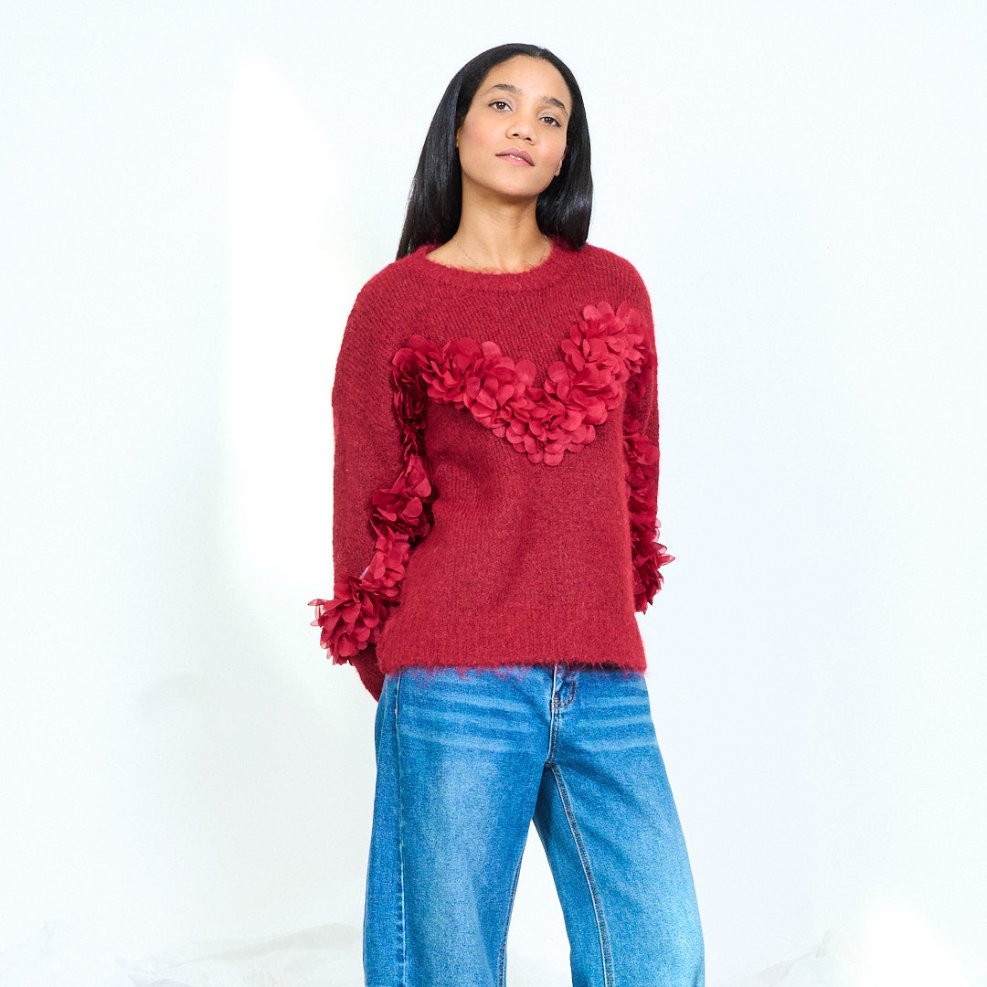 Model wearing a burgundy round-neck knit sweater with layered flower appliques across the chest and sleeves, front view