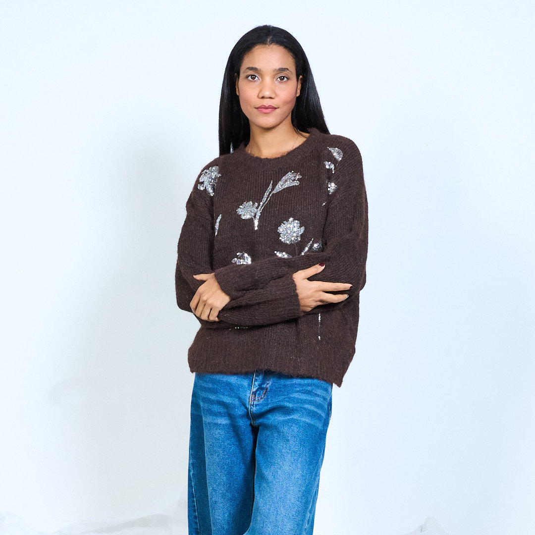 Model wearing a dark brown round-neck knit sweater with shiny flower and leaf embellishments, front view with folded arms