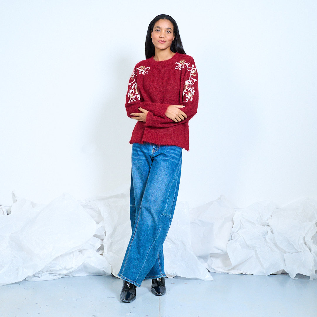 Model wearing a burgundy round-neck knit sweater with floral embroidery and shiny embellishments on the shoulders and sleeves, full-length front view