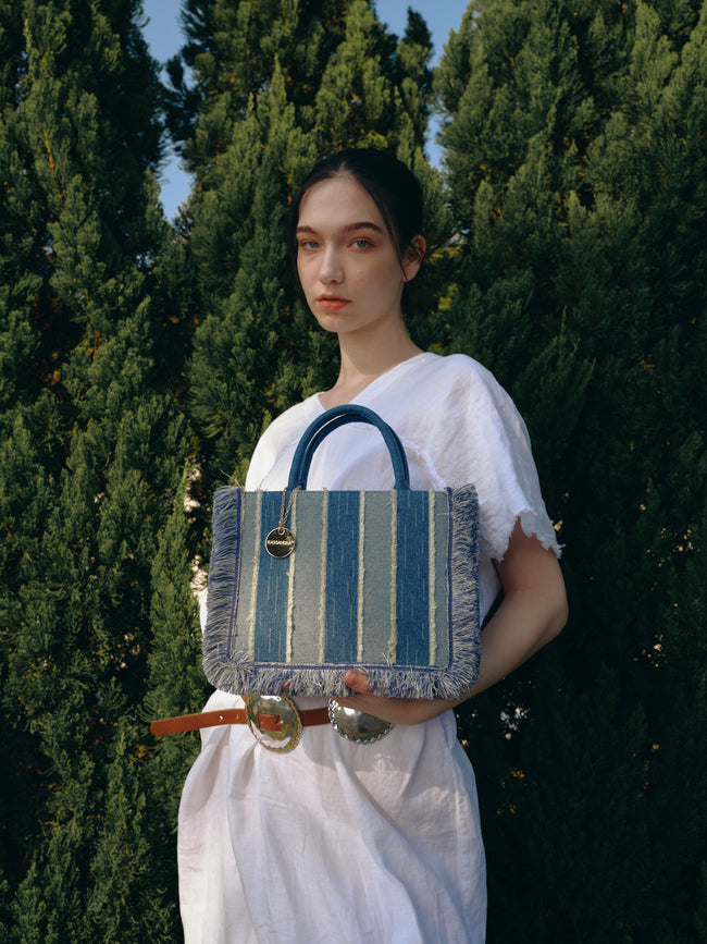 Model holding a rectangular blue striped tote bag with fringed edges and rounded handles outdoors.