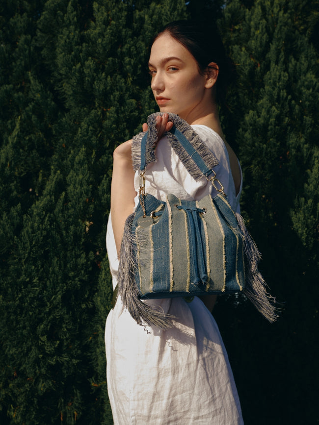 Model carrying a blue striped drawstring bucket bag with long side fringe and a fringed handle outdoors.