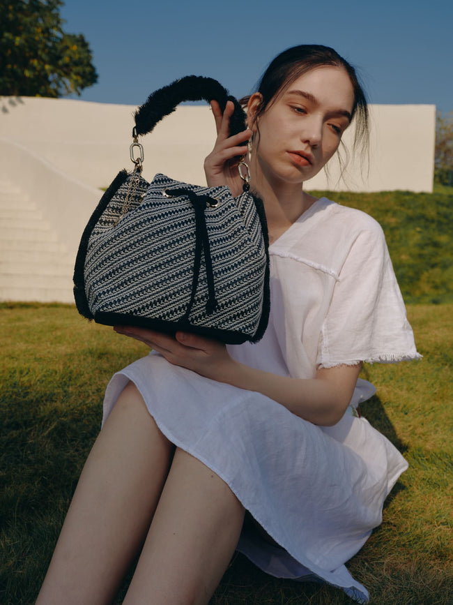 Seated model holding a black and white patterned bucket bag with a fringed handle and drawstring closure.