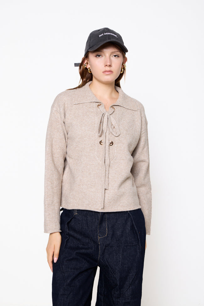 Beige taupe collared knit sweater with neck ties threaded through gold-tone eyelets, long sleeves and straight hem; jeans, cap and earrings styled separately