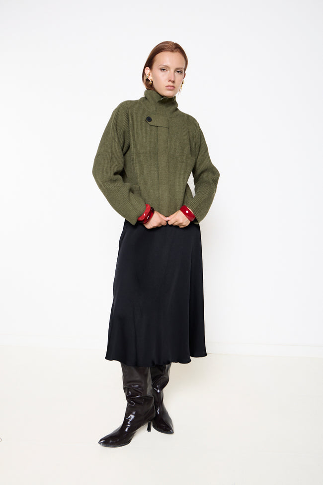 Dark olive green high-neck ribbed knit sweater with asymmetric chest button-tab detail, lower button details and long sleeves; skirt or jeans and earrings styled separately