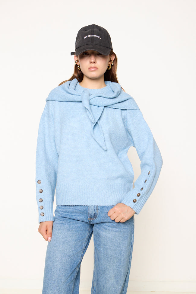 Light blue relaxed knit sweater photographed with matching scarf-style collar and front tie, decorative cuff buttons and ribbed hem; jeans, cap and earrings styled separately